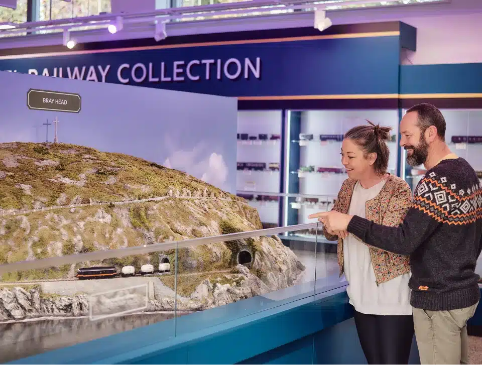 A man and woman smile and point while looking at a detailed miniature model of a hilly landscape with trains, displayed at a "Railway Collection." The scene includes a small tunnel and various terrain features.