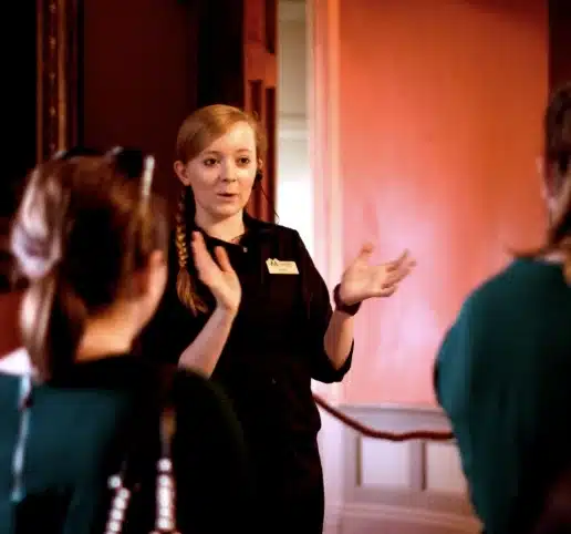 A woman with a braided hairstyle is passionately speaking and gesturing in front of a group, her name tag catching the light. Indoors, against a backdrop of Malahide Castle’s iconic red wall and an ajar door, she leaves her audience captivated by her presence.