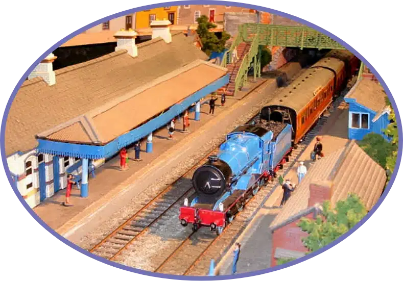 A blue toy train on a model railway with a vintage-style station. Minifigures stand on the platform. A bridge and buildings are in the background. The scene is enclosed in an oval frame.