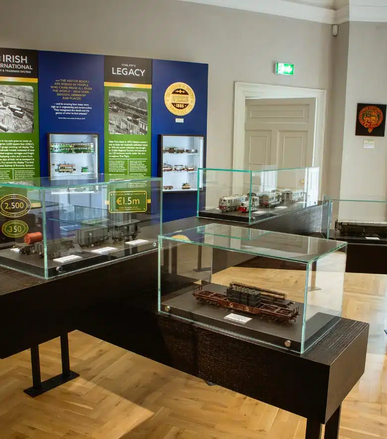 In a room reminiscent of Malahide Castle's charm, model trains are displayed in elegant glass cases on black stands. Blue panels on the back wall recount the history and legacy of Irish railways, while the hardwood floor adds warmth to this evocative exhibition space.