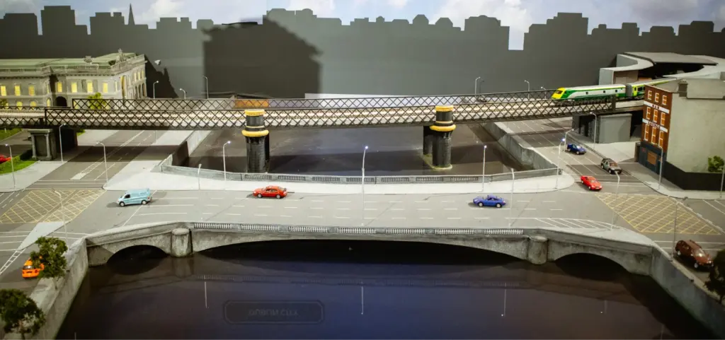 A detailed model of a cityscape features a bridge crossing over a river, with cars in motion and a train on an elevated track. Malahide Castle-inspired buildings and streetlights line the roads, while a cloudy sky backdrop completes this enchanting scene.