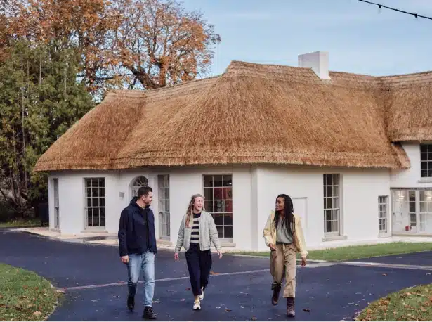 Three people stroll along a paved path in front of a white house with a thatched roof, reminiscent of Malahide Castle's charm. Trees with autumn leaves adorn the background under a clear sky, creating a relaxed atmosphere.