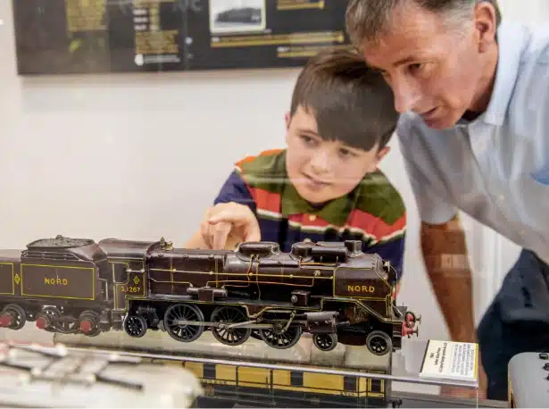 A boy and a man look intently at a detailed model train displayed in a glass case. The boy is pointing at the train, which is labeled "NORD" and "3.167". They appear to be in a museum or exhibition.