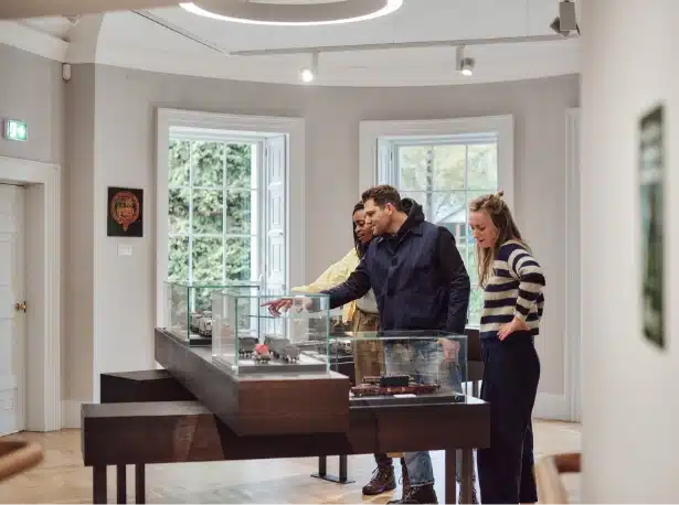 Three people in a gallery or museum observe display cases with objects inside. The room has large windows letting in natural light and a circular overhead light fixture. Walls are light-colored with minimal decorations.