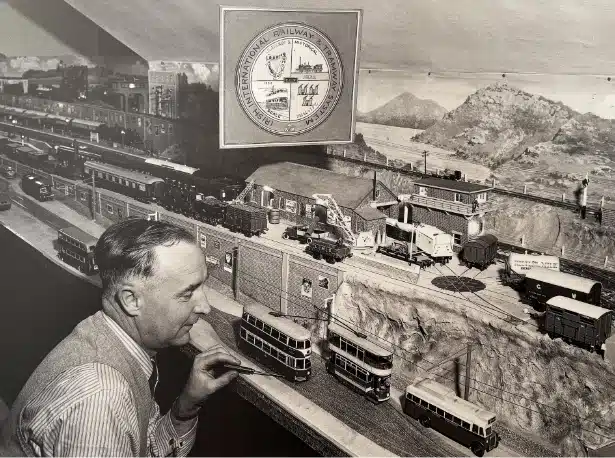 Man observing a detailed model train set with miniature trains, tracks, and landscapes including hills. A crest reading "National Railway... Festival" is visible above. The scene captures a vintage train enthusiast setting.