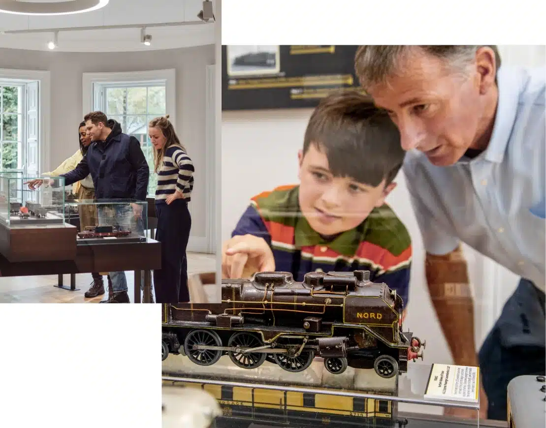 People in a museum are focused on model train exhibits. A young boy and an older man closely observe one train model, while two others in the background examine different displays. The room is well-lit with large windows.