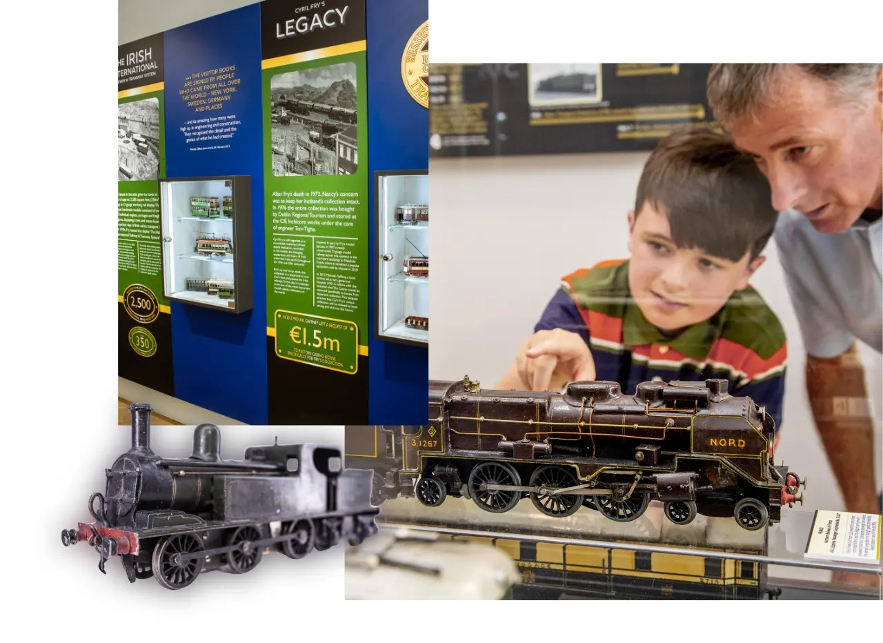 A boy and an older man look at model trains. To the left, a museum exhibit displays information panels and cases with historic Irish railway artifacts. Two model trains are shown at the bottom.