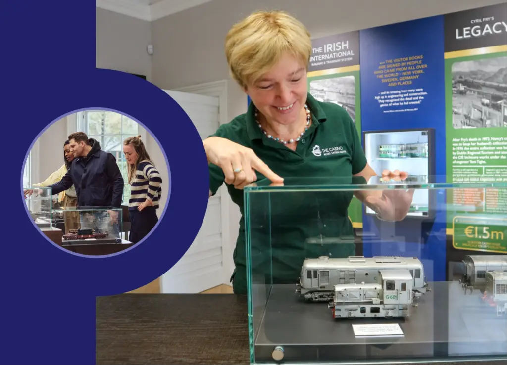 A woman in a green shirt points at a small train model inside a glass case at a museum. Inset shows three people observing another model. Exhibition boards with text and images are in the background.