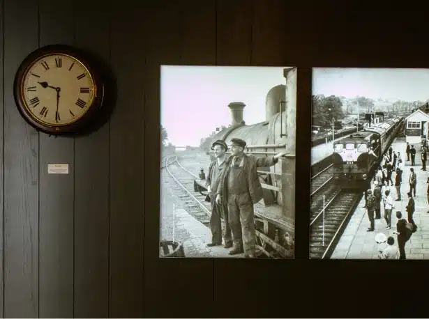 Wall-mounted vintage clock next to two black-and-white photos. One shows two men in 19th-century attire by a steam train, and the other depicts a busy train station platform with a departing train and people in period dress.