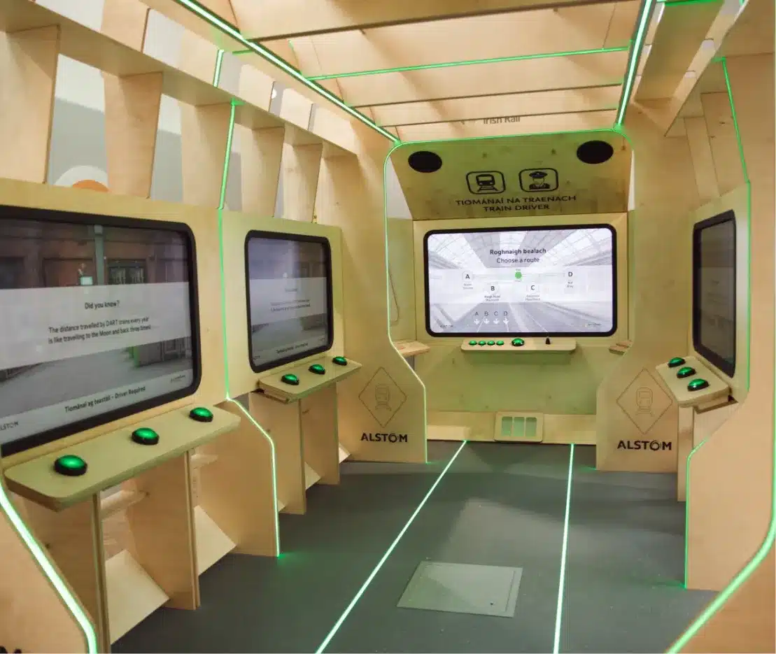 Interactive train simulator setup with wooden panels and green lighting. Multiple screens display digital interfaces, providing a realistic train operating experience. The floor and walls feature structured, modern designs, with "ALSTOM" branding visible.