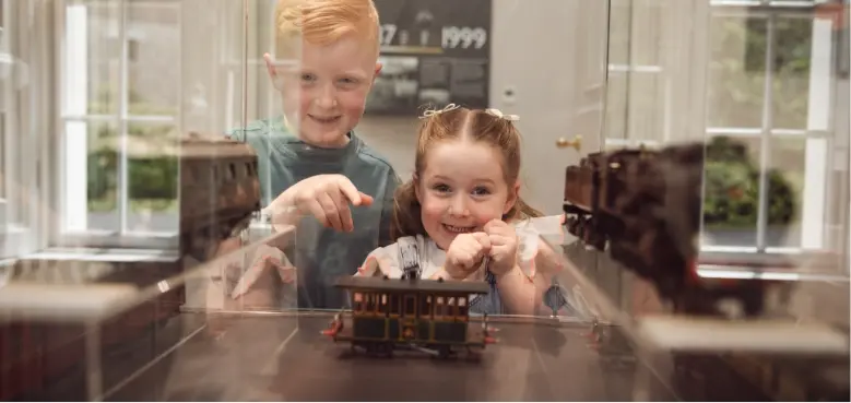 Two children excitedly point at a miniature train model encased in glass. They appear enthusiastic and engaged, with a blurry background of a room displaying other train models.