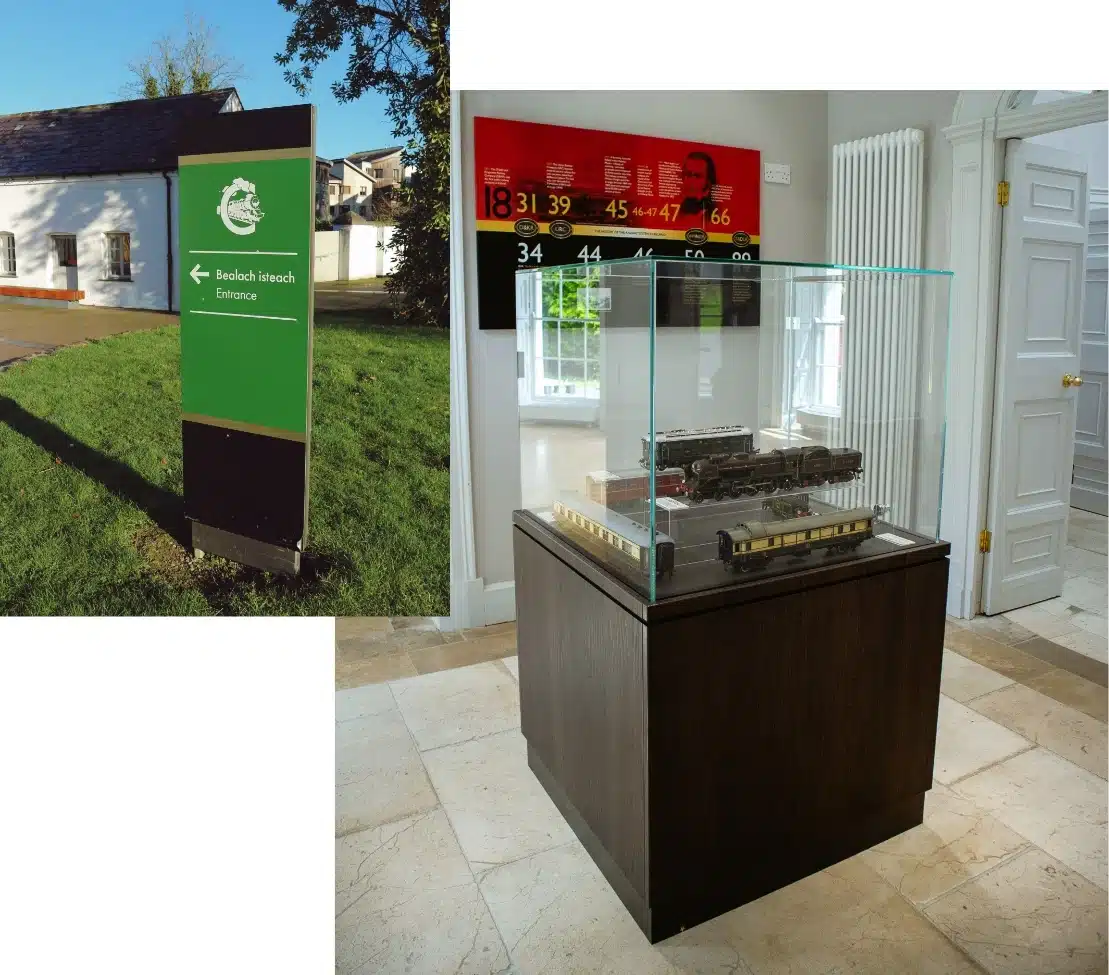 A museum display with a glass case showcasing a model train on a wooden base indoors. Adjacent is a green entrance sign with the Irish text "Bealach Isteach" outside, placed on grass near a white building.