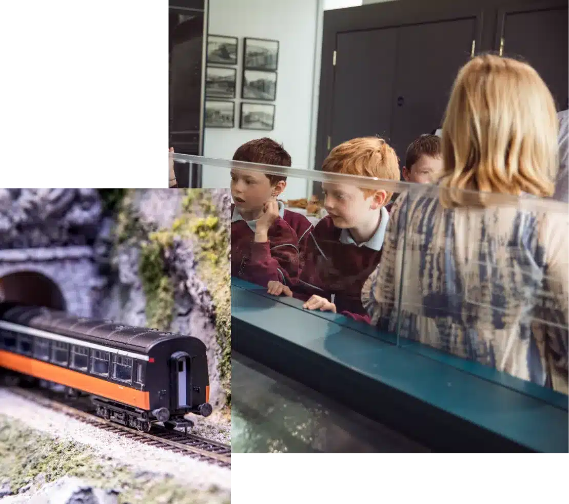 Children excitedly look at a model train set in a glass display. The train is black and orange with a realistic backdrop featuring a tunnel and rocky terrain. An adult is standing nearby with their back to the camera.