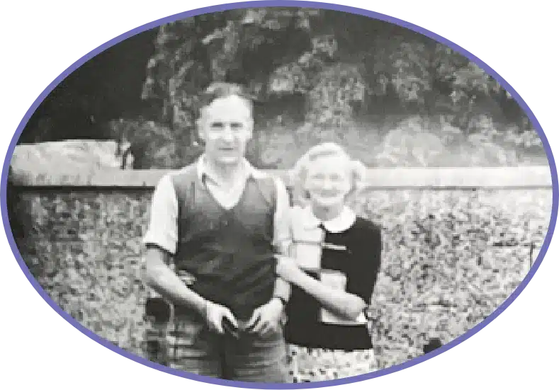 A black and white photo of a man and woman standing closely together, framed in an oval. They are outside, with a stone wall and trees in the background. The woman holds a book and smiles, while the man stands with his hand in his pocket.