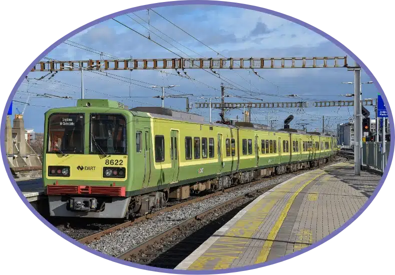 A green and yellow DART train travels along the tracks at a station, under clear blue skies. The tracks run parallel to a platform with yellow safety lines. Overhead power lines are visible against the sky.