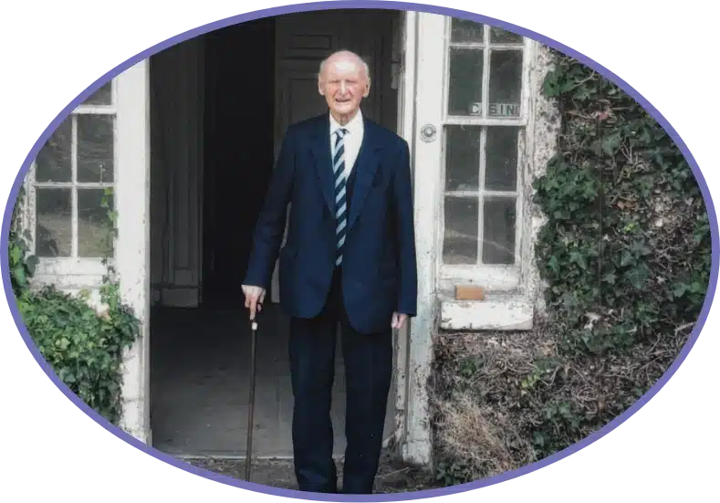 An elderly man in a suit with a blue striped tie stands holding a cane at the ivy-covered doorway of Malahide Castle. He's smiling, and the photo has an oval border.