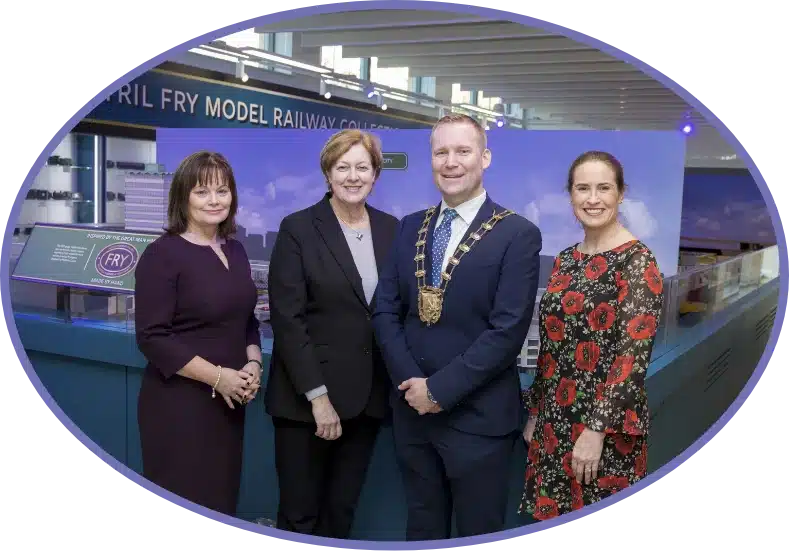 Four people are standing indoors, smiling at the camera. One person wears a chain of office, and the others don business attire. They pose in front of a display titled "Fry Model Railway Collection," located within the charming environment of Malahide Castle.