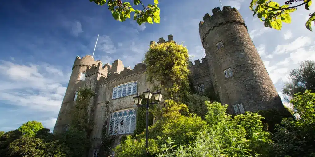 Malahide Castle, with its round towers and ivy-covered walls, stands majestically under a bright blue sky. Ornate windows peek through lush greenery, while a vintage lamp post graces the foreground.