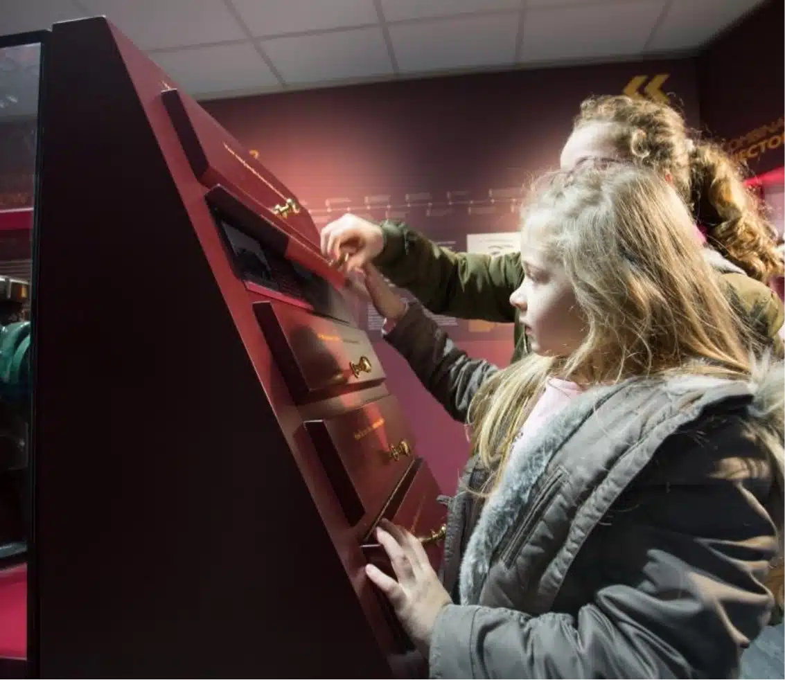 Two children interact with a museum exhibit consisting of a chest of drawers with small brass handles. The room is dimly lit, creating a focused atmosphere as they explore the contents. Both kids are wearing warm jackets.