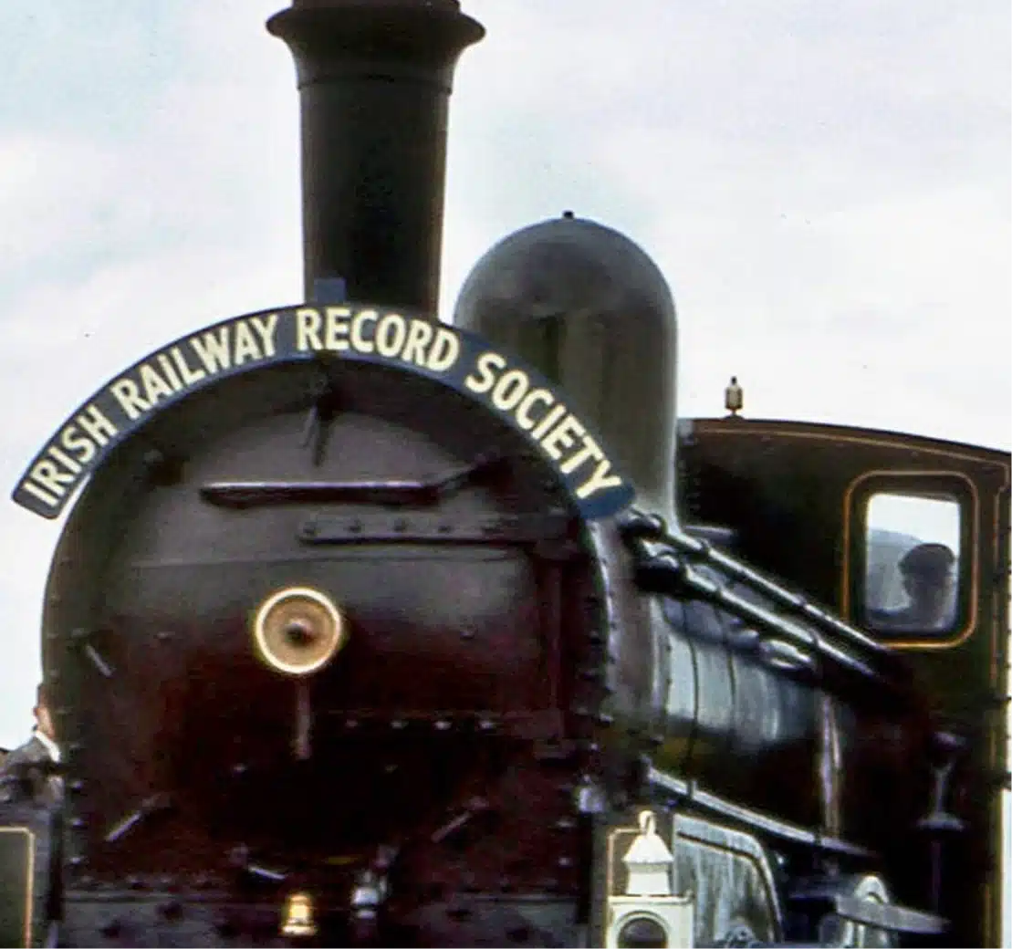 A vintage steam locomotive with a sign reading "Irish Railway Record Society" at the front. A person is visible inside the cabin. The locomotive is black with brass details. The sky is overcast.