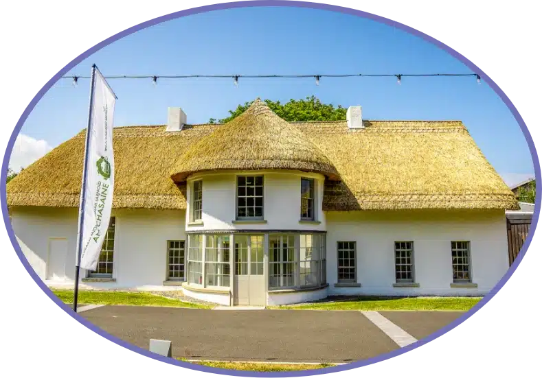 A white, round building with a thatched roof and multiple windows under a clear blue sky. A flag with green and purple text is in front, and there are string lights above. The photo is framed in an oval shape.
